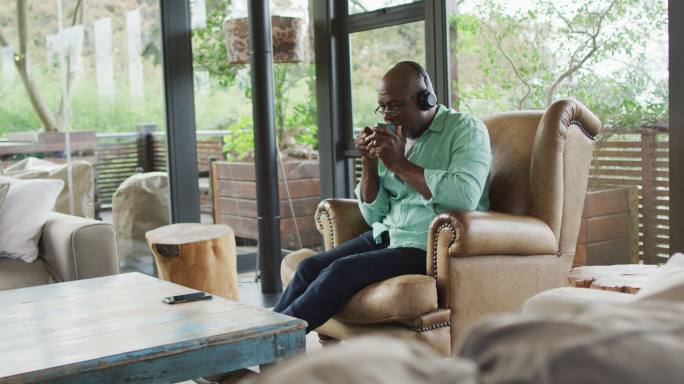 快乐的非洲裔美国老人放松在扶手椅，戴着耳机和喝咖啡