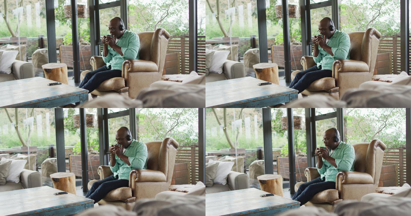 快乐的非洲裔美国老人放松在扶手椅，戴着耳机和喝咖啡