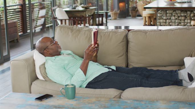 快乐的非裔美国老人放松，躺在沙发上看书