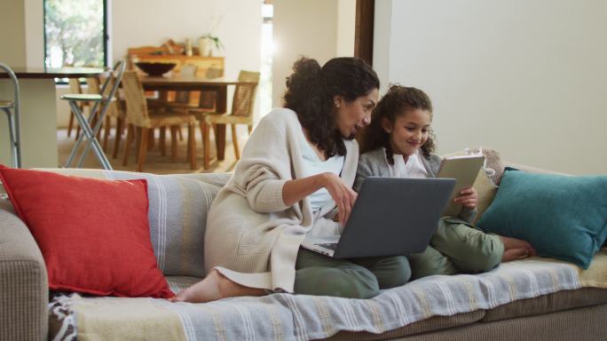 幸福的混血母亲和女儿坐在沙发上，玩得开心，并使用笔记本电脑