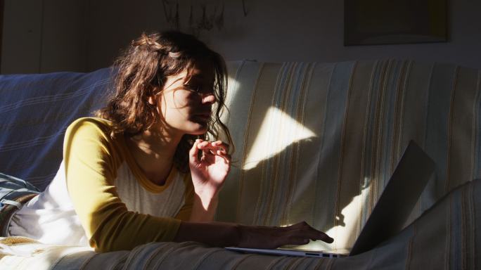 快乐的混血儿女人躺在沙发上使用笔记本电脑在阳光明媚的客厅微笑