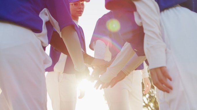 一群不同的女棒球运动员在阳光明媚的球场上，挤在一起，双手叠在一起