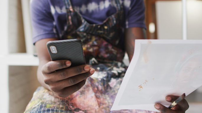 非裔美国男性画家在艺术家工作室里拿着智能手机和照片的中段
