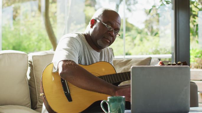 快乐的非洲裔美国老人在客厅玩吉他和使用笔记本电脑