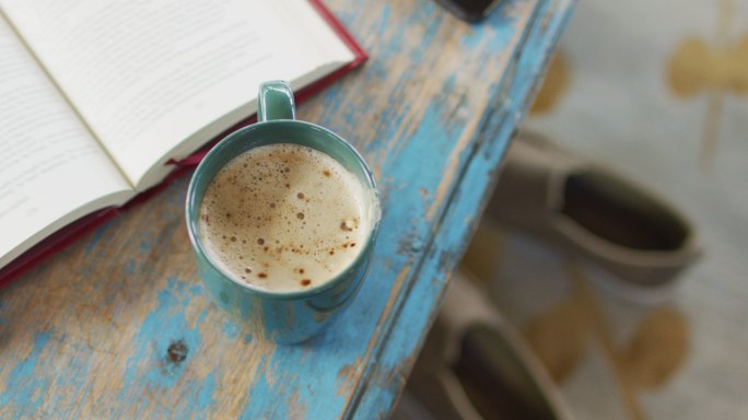 破旧的、涂过漆的木制桌面上放着笔记本和一杯加牛奶的咖啡，下面是地板上的拖鞋