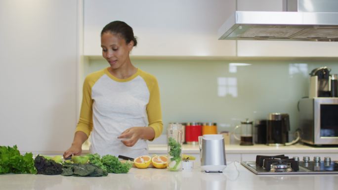 混合种族妇女准备健康的饮料，切水果和蔬菜在厨房