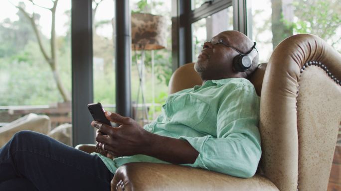 快乐的非洲裔美国老人放松在扶手椅，听使用耳机和智能手机