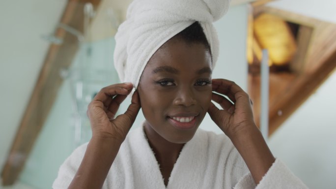 微笑的非洲裔美国人迷人的女人把毛巾放在头上，在浴室里看着镜子