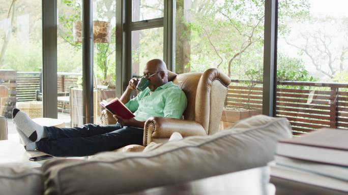 快乐的非洲裔美国老人放松在扶手椅，脚，看书和喝咖啡