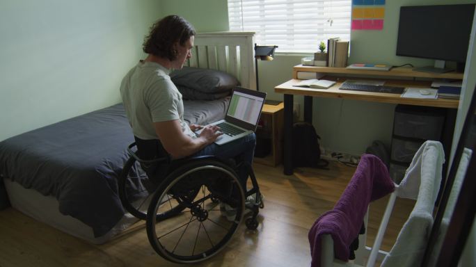 重点关注坐在轮椅上的白人残疾人在卧室里使用笔记本电脑