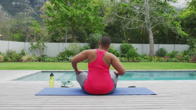 快乐的非裔美国胖女人练习瑜伽，在花园里的游泳池边伸展身体