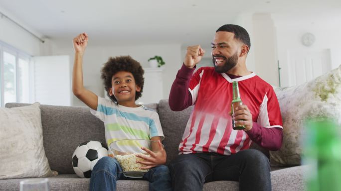 非洲裔美国人和他的儿子坐在家里客厅的沙发上，看电视上的体育节目