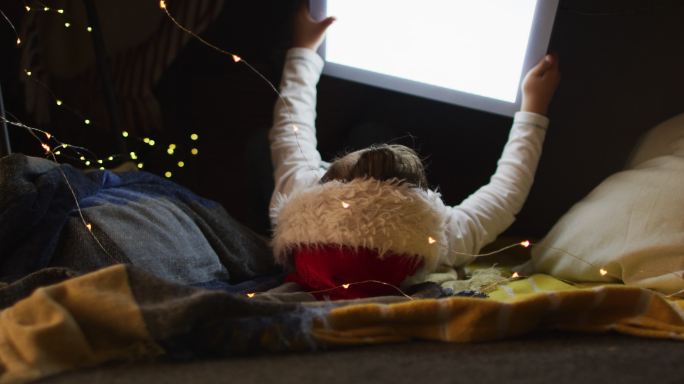 白人男孩戴着圣诞老人帽，躺在枕头上玩平板电脑