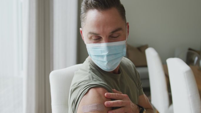一名白人男子戴着口罩，在家里展示他接种过疫苗的肩膀