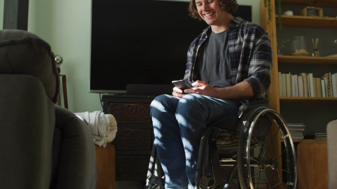 快乐的白人残疾人坐在轮椅上在客厅里用智能手机