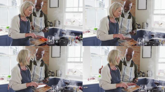 混合种族的老年夫妇穿着围裙，在家里的厨房里用数码平板电脑做饭