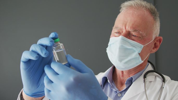 白种人男资深医生戴着口罩手持新冠疫苗