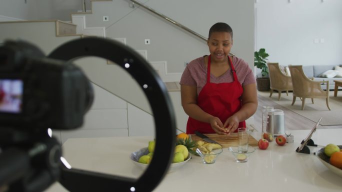 快乐的非裔美国胖女人穿着围裙，在厨房用相机制作烹饪视频博客