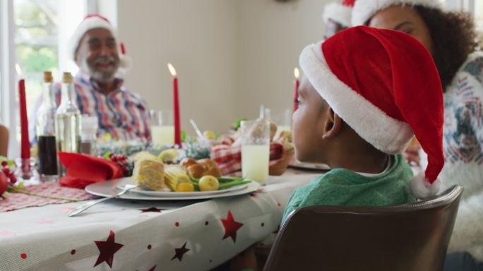 微笑的非裔美国男孩的肖像，戴着圣诞老人的帽子与家人庆祝节日大餐