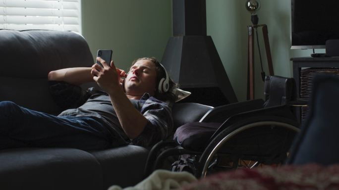放松的白人残疾人躺在客厅的沙发上，戴着耳机，使用智能手机