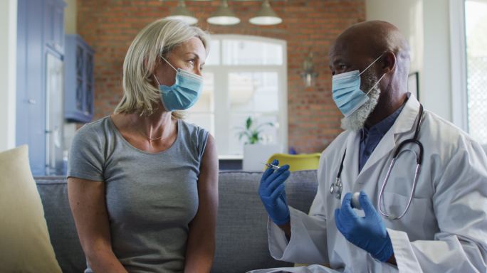 快乐的高级多样化的女人和医生戴着口罩坐在客厅的沙发上，接种疫苗