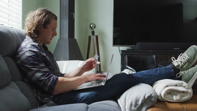 快乐的白种人坐在客厅的沙发上，拿着卡片，用着笔记本电脑