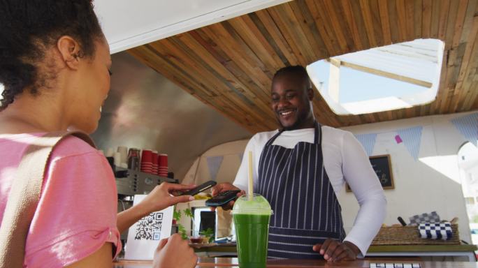 快乐的女性顾客用智能手机付钱给微笑的非洲裔美国男性餐车老板
