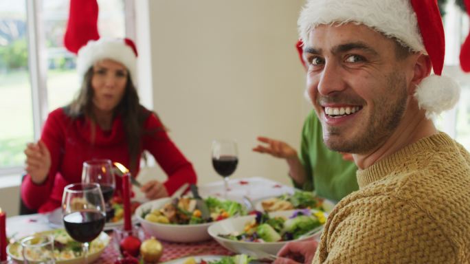 快乐的白人男子戴着圣诞帽看着相机在圣诞大餐