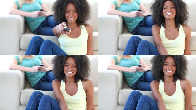 一名黑人妇女在看电视，而她的朋友正在沙发上玩手机