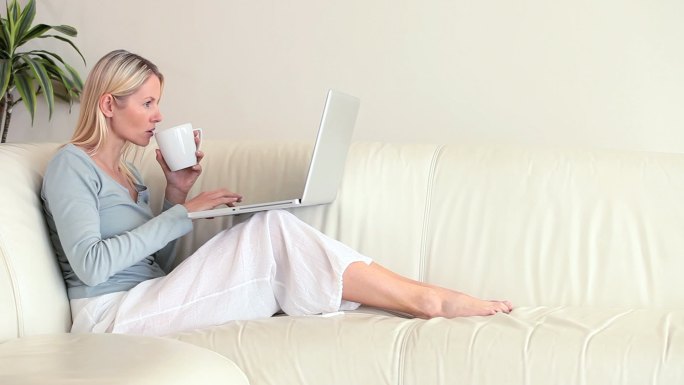 一个白人女人坐在沙发边看笔记本电脑边喝水