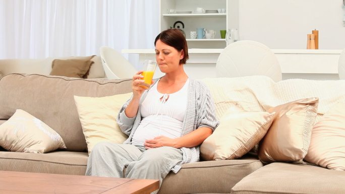 深色头发的孕妇坐在沙发上喝着橙汁
