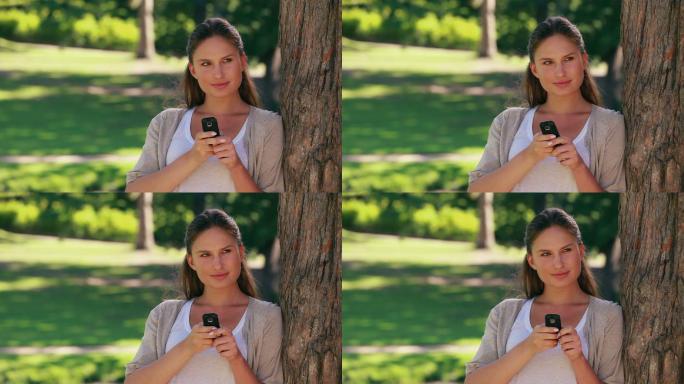 在公园里，一名妇女靠在一棵树上，手里拿着手机