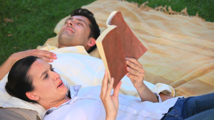一个女人在给她躺在公园草地上的丈夫读书