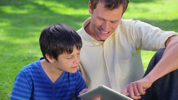 一名微笑的男子和他的儿子在公园里使用平板电脑