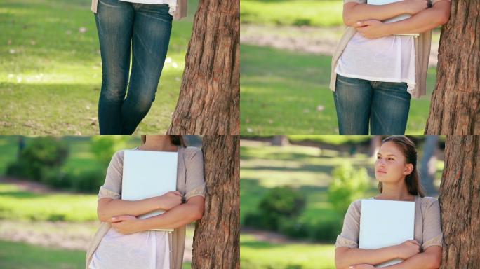 一个女人拿着文件，倚靠在公园的一棵树上