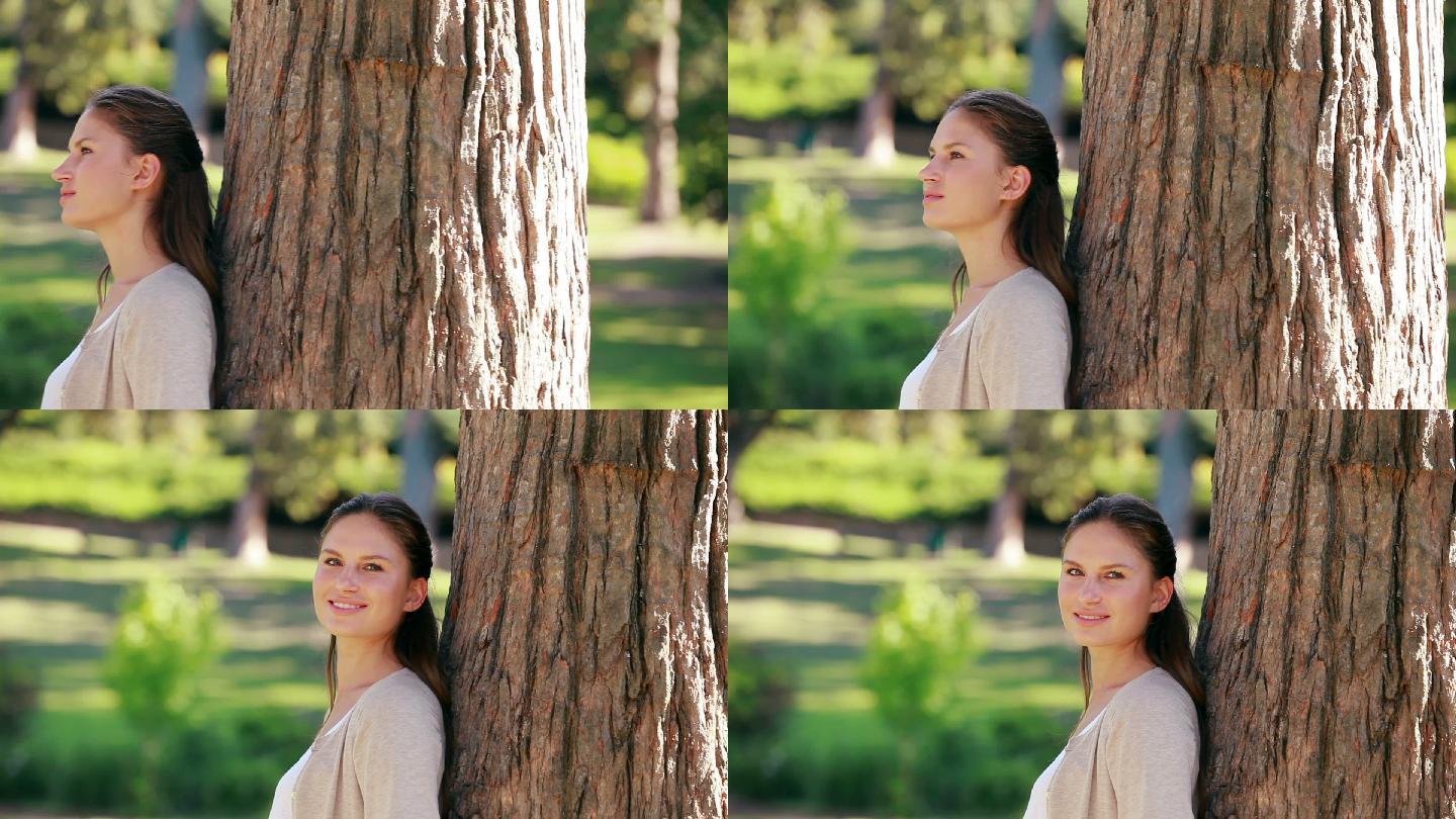 一个微笑的女人，靠在公园的一棵树上