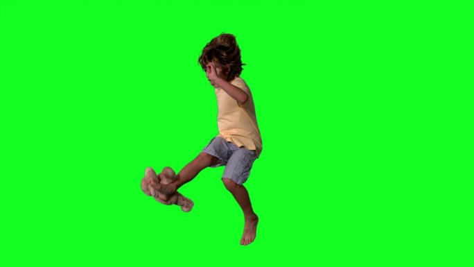 在慢镜头中，一个小男孩在绿色屏幕上跳起来踢泰迪熊