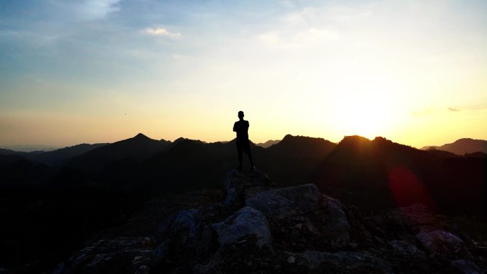 男人抱手站山顶眺望战胜自己夕阳逆光剪影