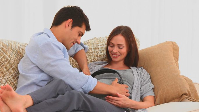 一名男子在起居室里把耳机戴在他怀孕的妻子的肚子上