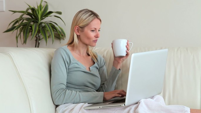 一个女人坐在沙发上，一边用笔记本电脑打字，一边拿着杯子喝水