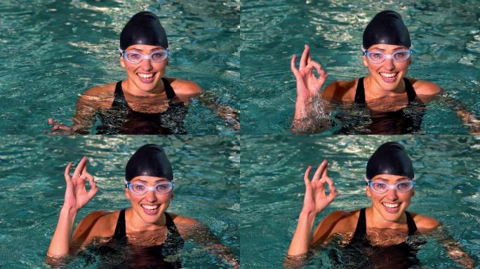健康的女游泳运动员在游泳池慢动作显示ok的迹象