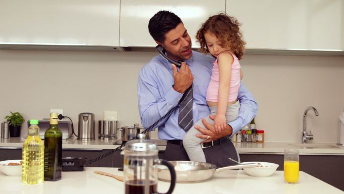 微笑的父亲抱着他的小女儿在家里的厨房打电话