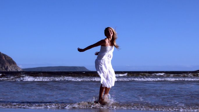 在阳光明媚的日子里，美丽的金发女郎穿着白色连衣裙在沙滩上跳起了慢镜头