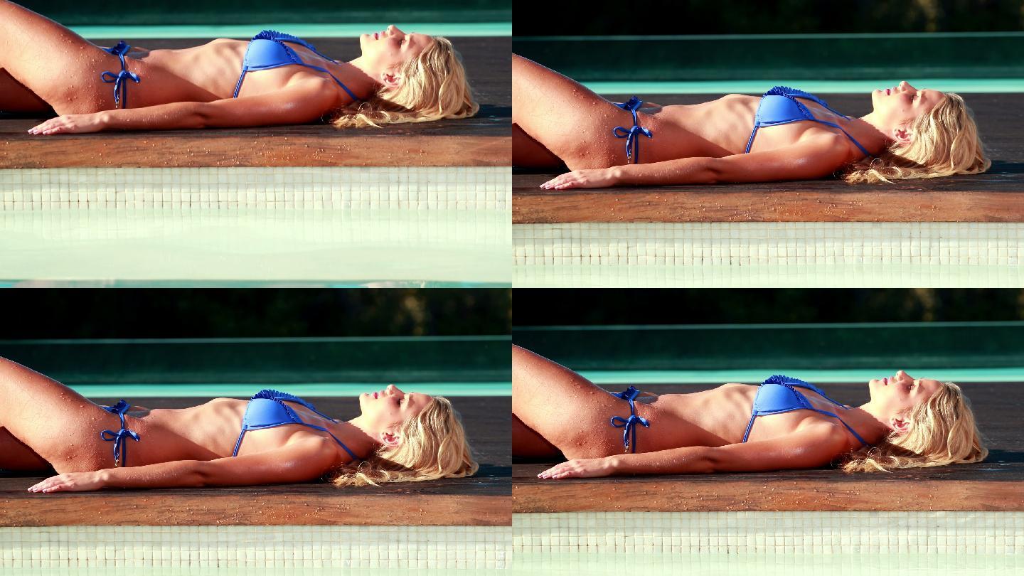 性感的金发女郎在蓝色比基尼放松躺在一个阳光明媚的日子的泳池边