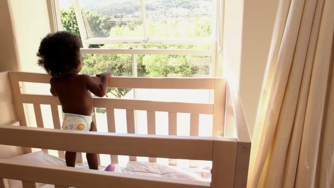 可爱的小女孩站在她的婴儿床上望着窗外，在家里的卧室