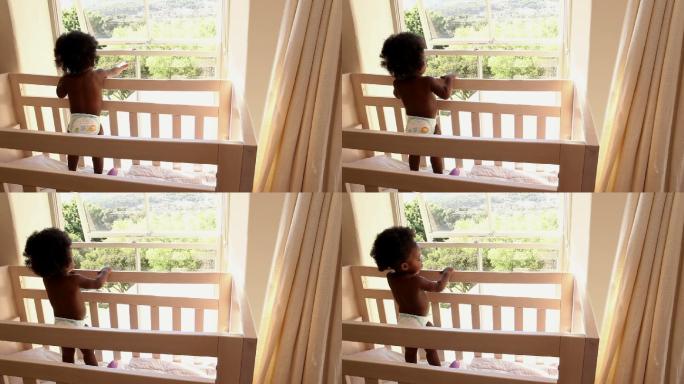 可爱的小女孩站在她的婴儿床上望着窗外，在家里的卧室