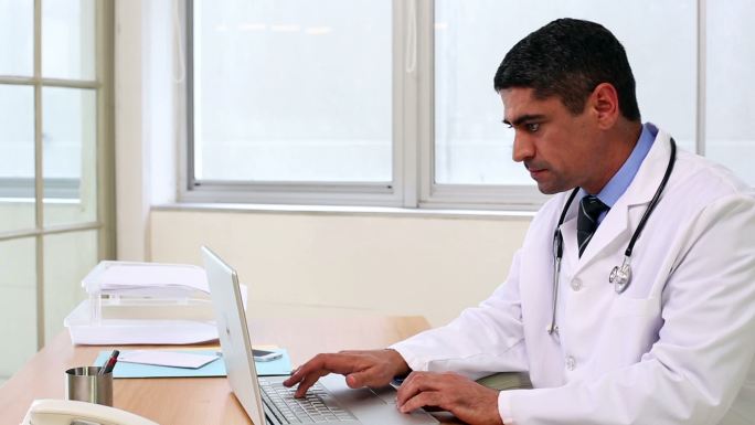 医生坐在桌子上用笔记本电脑打字在医院的办公室里