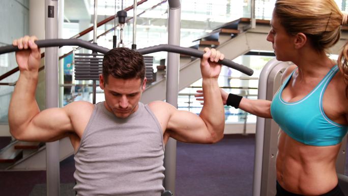 健身的人在健身房用举重机锻炼手臂，而教练在监督