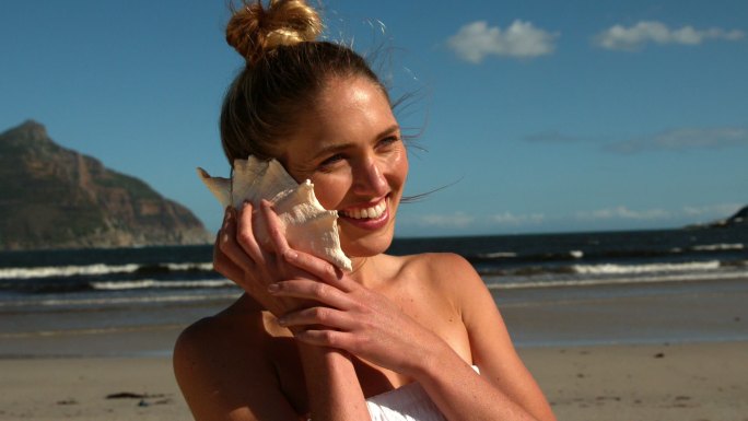 微笑的金发女郎听着海滩上海螺的慢镜头