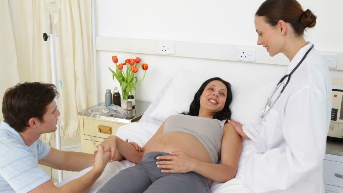 黑发孕妇躺在床上与伴侣和医生在医院谈话